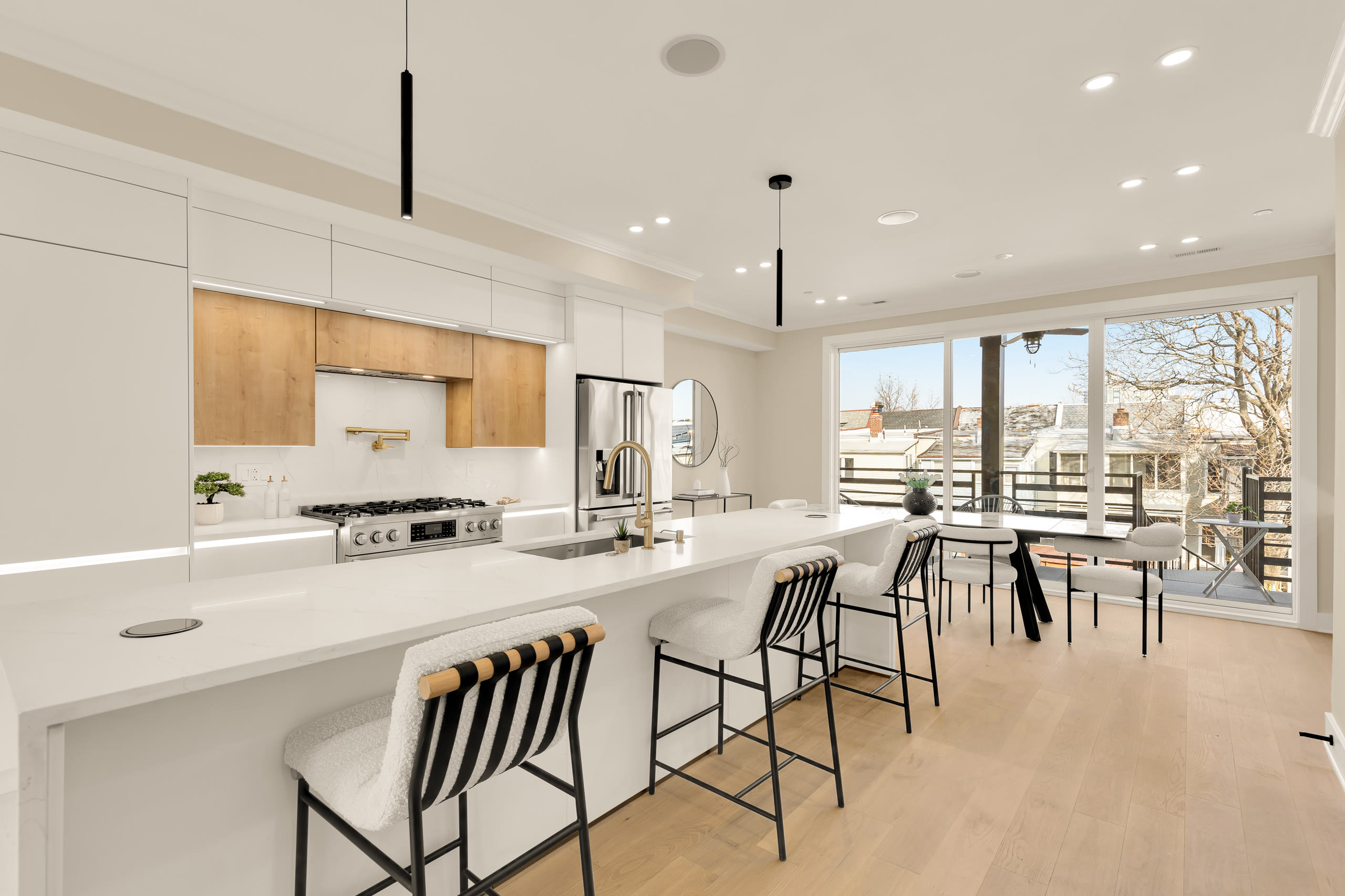 White flat-front kitchen with wood upper cabinets above the range, a white quartz waterfall island, and pendant lights opening to a dining area.