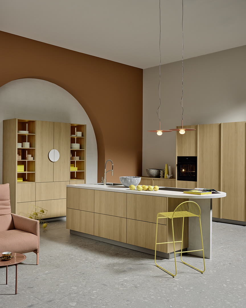 Light wood kitchen with a white waterfall island, handle-free drawers, and open shelving flanking an arched opening.