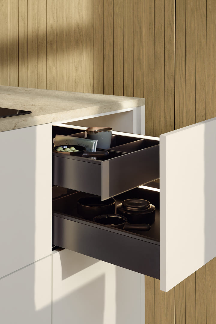 Open kitchen drawer with white fronts and black interior organizers beside a light stone countertop