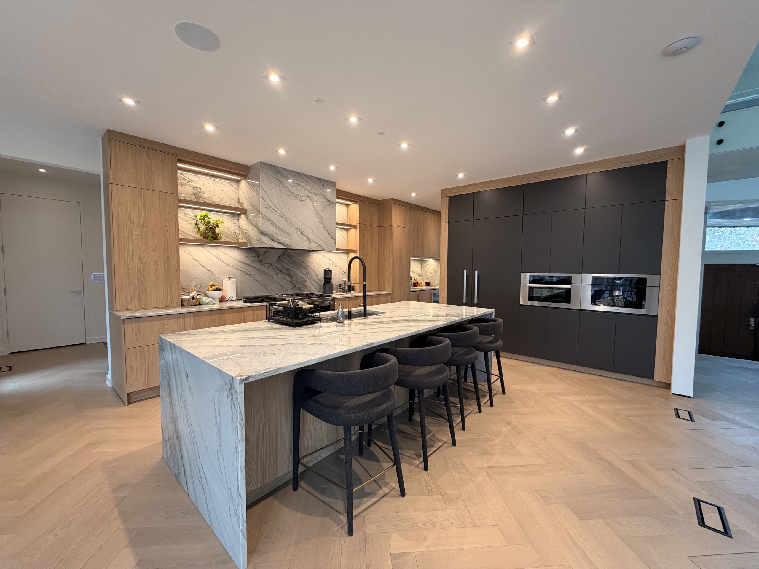 Finished kitchen with light wood cabinets, a marble waterfall island with bar stools, and dark handleless wall units