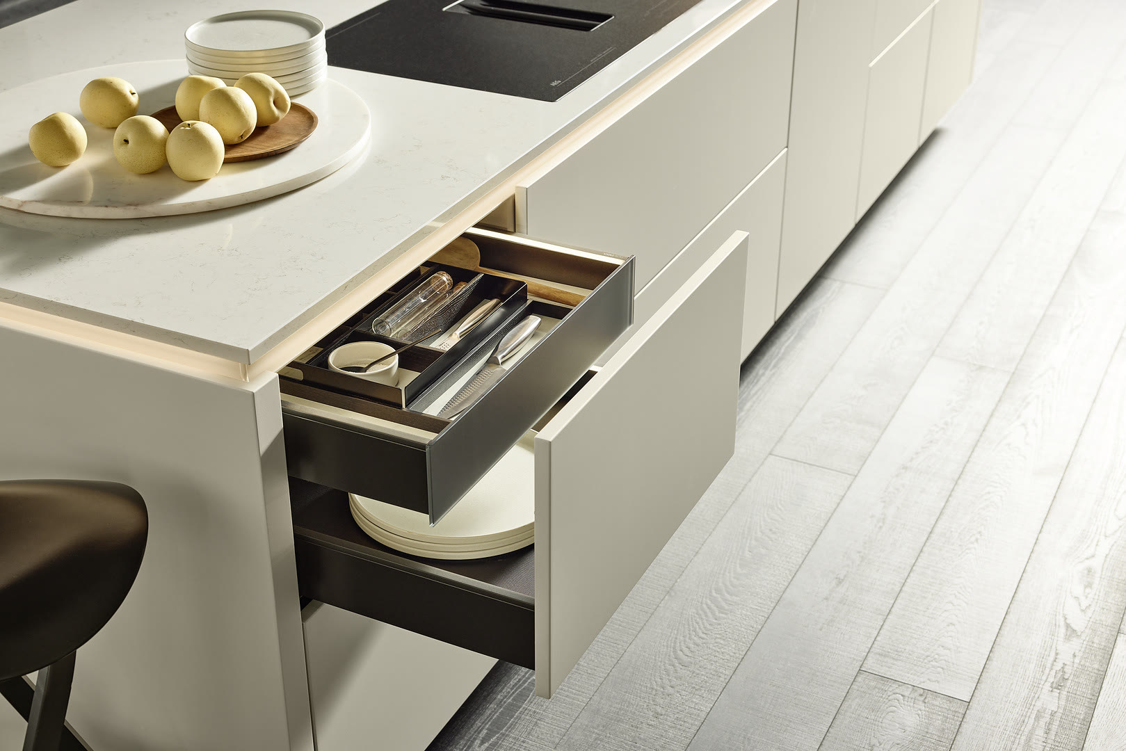 Open kitchen island drawer with utensil organizers and dishware beside a black induction cooktop