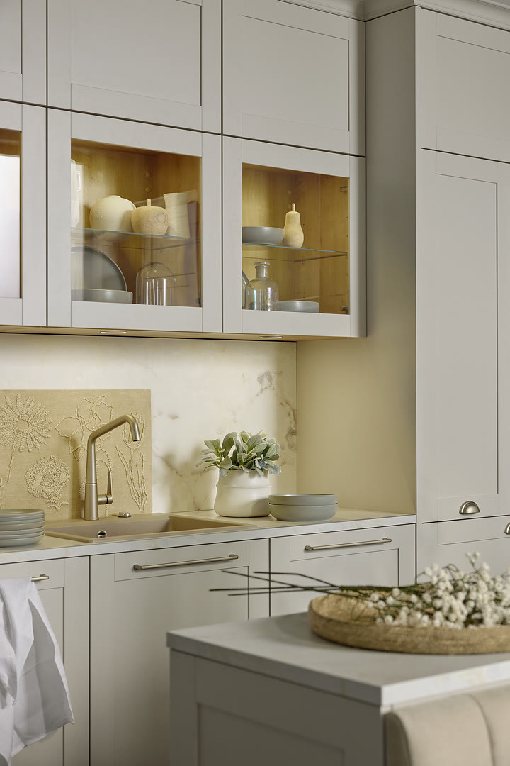 A finished kitchen with white shaker-style cabinetry, glass-front upper cabinets with brass interior backing, cream-coloured countertops
